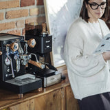 A woman in glasses and a cream sweater reads a magazine beside a black Lelit Mara X3 Semi-Automatic Heat-Exchange Espresso Machine PL62X3 and a coffee grinder on a wooden counter against a brick wall.