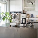 In a modern kitchen, sunlight illuminates a white Lelit Mara X3 Semi-Automatic Heat-Exchange Espresso Machine PL62X3 by Lelit, along with a coffee grinder, milk frother, and kettle on a gray countertop beside a green plant.