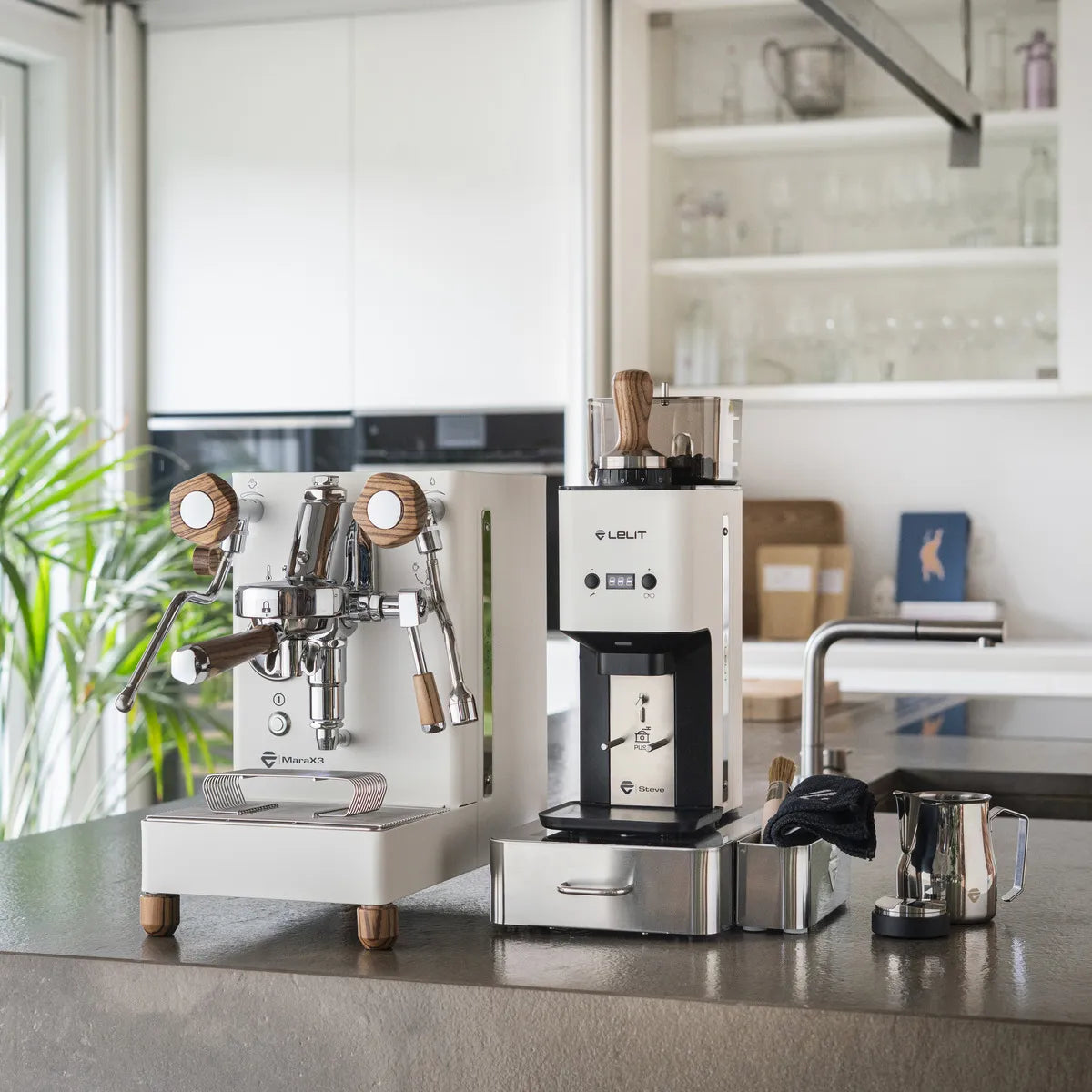 A modern kitchen features the Lelit Mara X3 Semi-Automatic Heat-Exchange Espresso Machine PL62X3 (White), coffee grinder, milk frothing pitcher, tamper, and cloth on the counter. Glassware and decor are visible in the background.