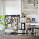 A modern kitchen features the Lelit Mara X3 Semi-Automatic Heat-Exchange Espresso Machine PL62X3 (White), coffee grinder, milk frothing pitcher, tamper, and cloth on the counter. Glassware and decor are visible in the background.