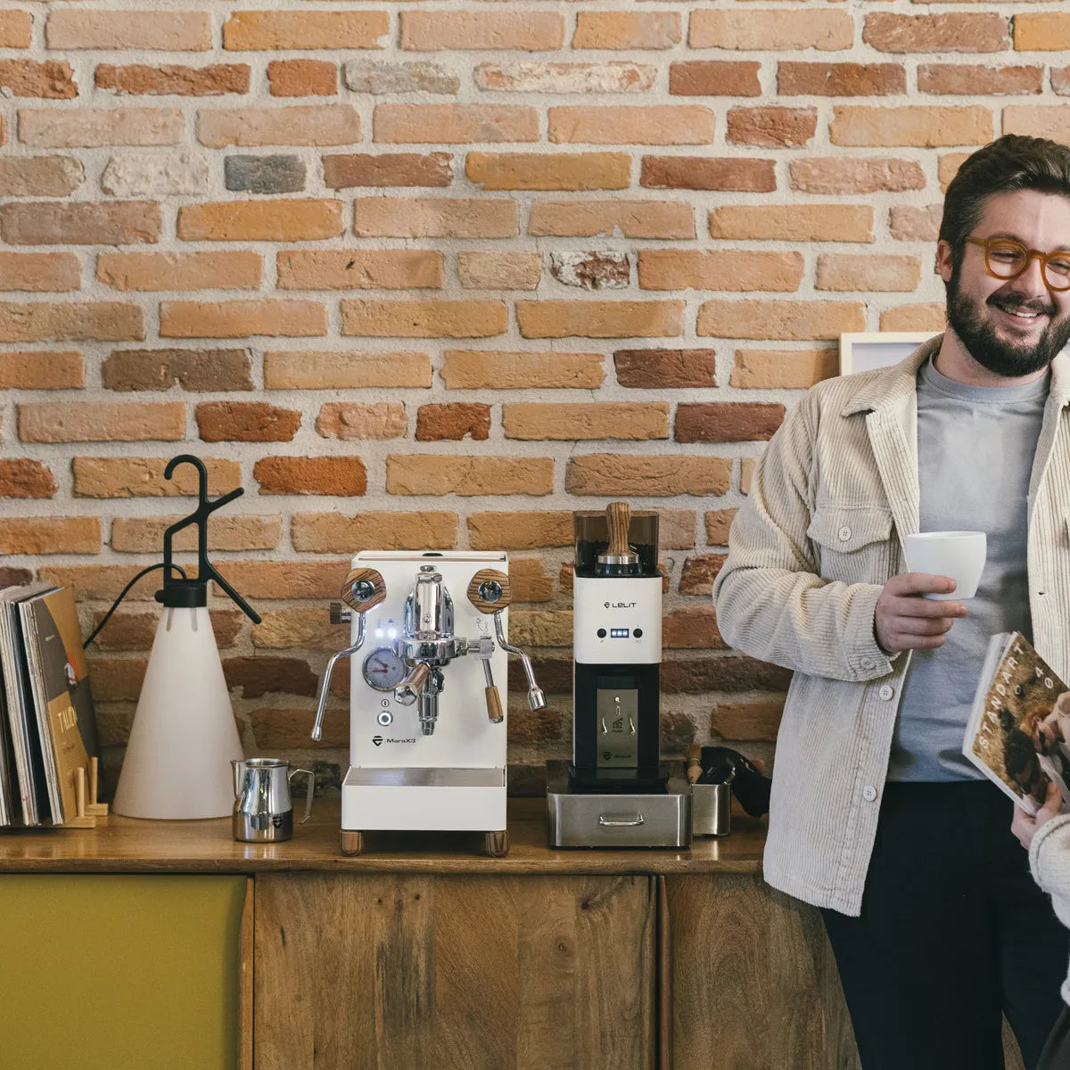 A man in glasses and a light jacket smiles while holding a cup beside a wooden counter with a Lelit Mara X3 Semi-Automatic Heat-Exchange Espresso Machine PL62X3 (White), grinder, mugs, and books against a brick wall background.
