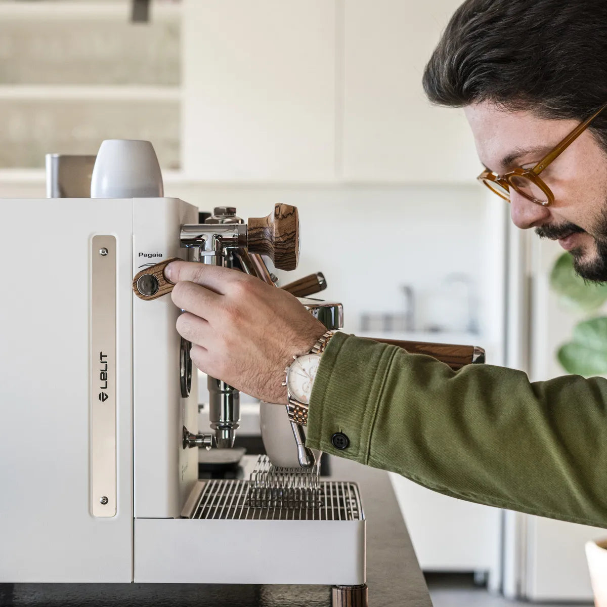 A man in glasses and a green shirt uses the Lelit Mara X3 Semi-Automatic Heat-Exchange Espresso Machine PL62X3 (White) to make coffee by pulling a lever in a modern kitchen.