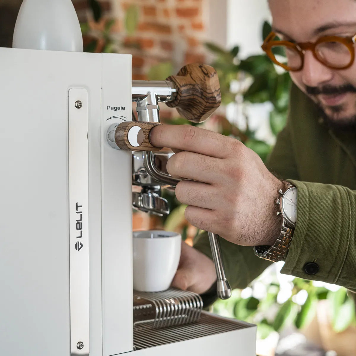 A person with glasses uses a white Lelit Mara X3 Semi-Automatic Heat-Exchange Espresso Machine (PL62X3), placing a portafilter and holding a cup underneath to make coffee. Green plants and a brick wall are seen in the background.