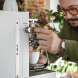 A person with glasses uses a white Lelit Mara X3 Semi-Automatic Heat-Exchange Espresso Machine (PL62X3), placing a portafilter and holding a cup underneath to make coffee. Green plants and a brick wall are seen in the background.