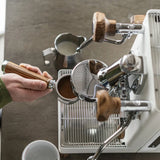 A person holds a portafilter with ground coffee, ready to use the Lelit Mara X3 Semi-Automatic Heat-Exchange Espresso Machine PL62X3 (White). A metal milk pitcher and empty cup from Lelit are on the countertop nearby.