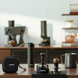 The MHW-3BOMBER Master Barista Set Bundle, featuring premium coffee tools, is showcased on a wooden counter with blurred machines and shelves in the background under warm, minimalist lighting.