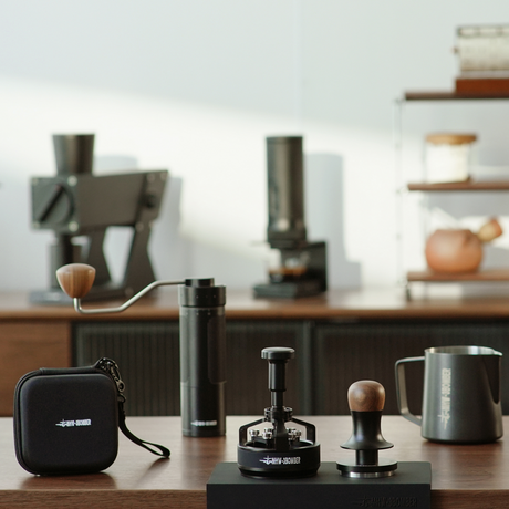 The MHW-3BOMBER Master Barista Set Bundle, featuring premium coffee tools, is showcased on a wooden counter with blurred machines and shelves in the background under warm, minimalist lighting.