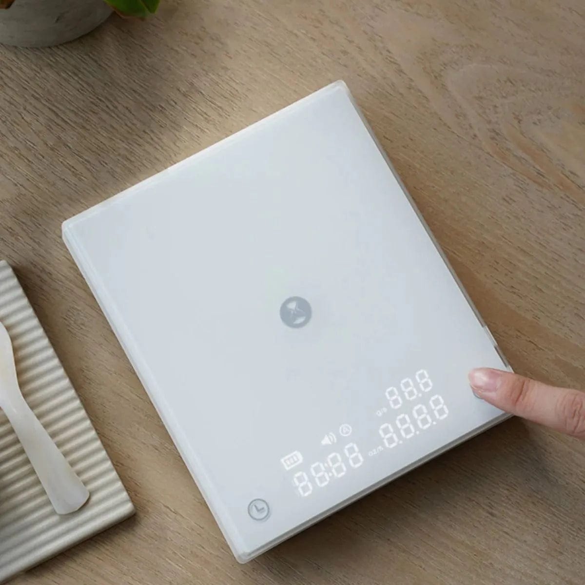 A digital kitchen scale is placed on a wooden table. The sleek, rectangular Timemore Mirror BASIC 2 Espresso Coffee Scale (White) by Timemore features a backlit LED display showing placeholder numbers. A person's finger touches the bottom right corner of this precision brewing scale, likely interacting with its controls.