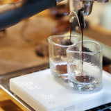 A close-up of a coffee machine portafilter dispensing espresso into two small transparent glasses, which are placed on a Timemore Mirror BASIC 2 Espresso Coffee Scale (White). The scale shows a timer and measurement reading, emphasizing precision brewing. The background is blurred, focusing attention on the brewing process.