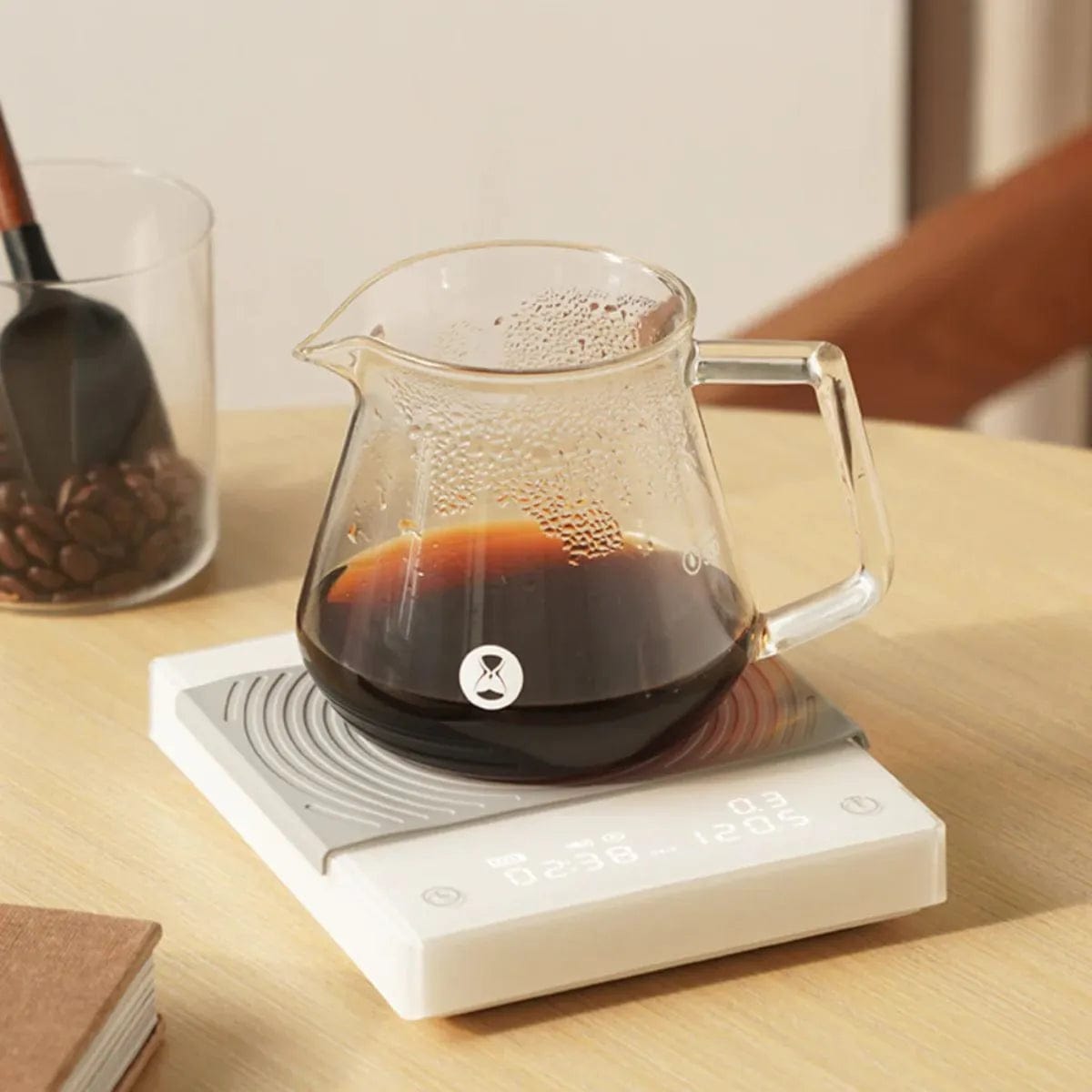 A clear glass jug containing dark coffee rests on a Timemore Mirror BASIC 2 Espresso Coffee Scale (White) atop a light wooden table. Nearby, a transparent container holds a black-handled scoop and coffee beans. A brown notebook lies in the foreground, perfectly set for precision brewing with flow rate monitoring.