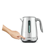 A hand presses a button on the Breville the Smart Kettle Luxe 1.7L BKE845BSS, a brushed stainless steel kettle with variable temperature control and water level window, set against a plain white background.