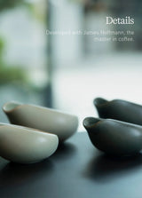 Two Loveramics Coffee Dosing Trays in matte dark green sit on a dark surface with a blurred background. Text reads: "Details. Developed with James Hoffmann, the master in coffee." A perfect complement to your coffee grinder accessory setup, these trays invite precision and enjoyment for every dose.