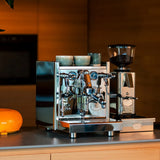 The ECM Technika VI Dual Boiler Espresso Machine w/ PID by ECM, topped with two cups, sits on a kitchen counter beside a coffee grinder filled with beans, set against warm wooden cabinetry.