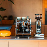 The ECM Technika VI Dual Boiler Espresso Machine w/ PID by ECM, with two cups on top, sits beside a coffee grinder filled with beans on the kitchen counter. A yellow lamp and plant appear in the background.