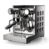 The Rocket Appartamento TCA Espresso Machine RE502A3B12 is a sleek, stainless steel premium domestic espresso machine with a modern design. It features a portafilter, pressure gauges, multiple adjustment knobs, and Temperature Control Adjustment. The "Rocket" brand logo is prominently displayed on the front of the Appartamento TCA, and circular cut-out patterns elegantly adorn the sides.