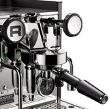 A close-up of the sleek Rocket Appartamento TCA Espresso Machine (RE502A3B12) in stainless steel with a black handle shows its chrome dials and "R" logo. Its polished industrial design highlights this premium domestic espresso machine featuring flow and temperature control adjustment.