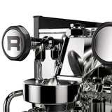 A close-up of the Rocket Appartamento TCA Espresso Machine (RE502A3B12) showcases its sleek stainless steel design with black accents. The round knobs and polished surfaces allow for precise temperature control, reflecting light off its modern, industrial look.