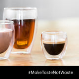 Three Bodum Pavina Small 2.5oz Double Walled Coffee & Espresso Glasses sit on a wooden table, each holding a different drink: tea, hot chocolate, and coffee. A hashtag at the bottom reads "#MakeTasteNotWaste.