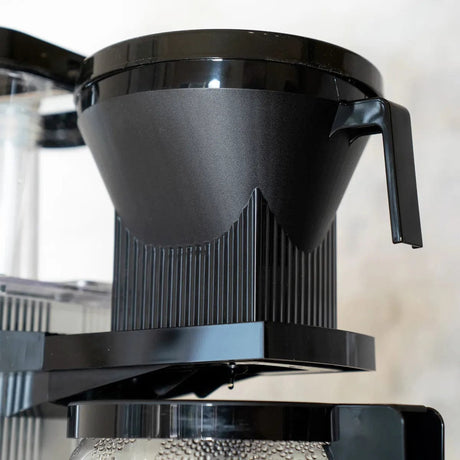 A close-up of the Moccamaster Technivorm Moccamaster Automatic Drip-Stop Brew Basket 13253. The ribbed, slightly conical filter is made from BPA-free plastics and features a side handle and a bottom spout for coffee to drip into a glass carafe placed below.