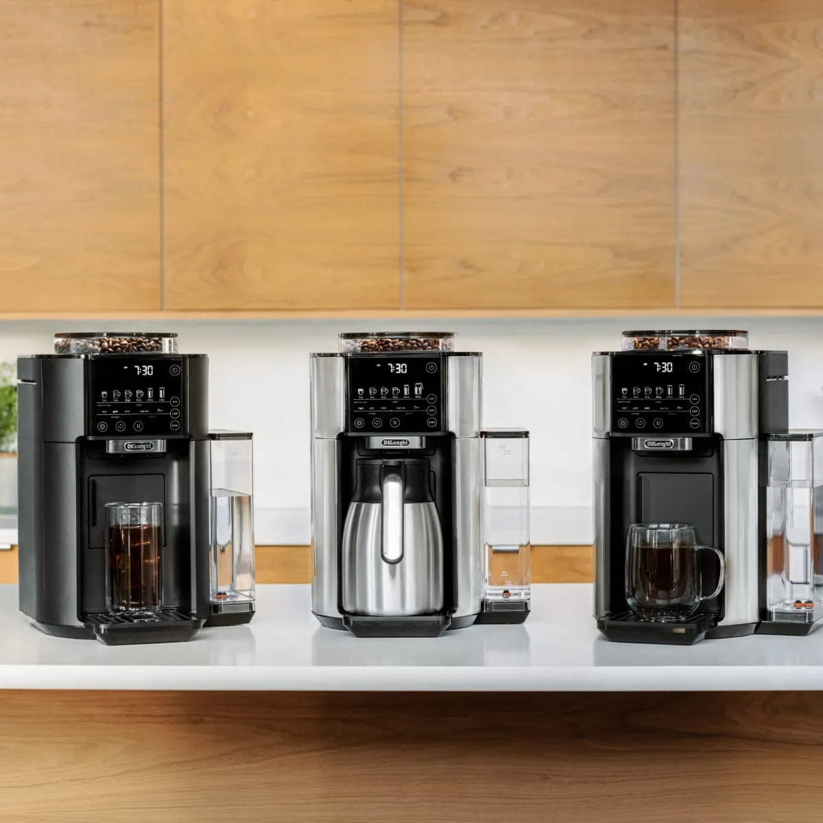 Three sleek DeLonghi TrueBrew Fully Automatic Drip Coffee Machines CAM51035M in stainless steel are arranged on a white countertop against light wood cabinets. Each coffee maker features a digital display and a transparent bean container on top. One machine dispenses coffee into its thermal carafe, while the other two serve single-serve drip coffee directly into mugs.