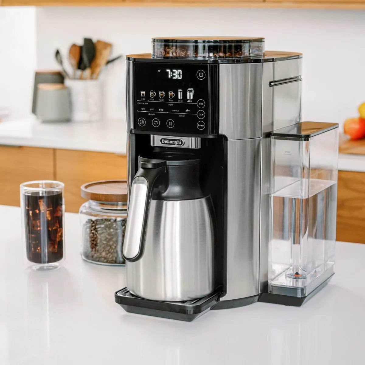 A sleek, modern DeLonghi TrueBrew Fully Automatic Drip Coffee Machine CAM51035M in stainless steel with a thermal carafe sits on a kitchen counter. It features a digital display showing 7:30, multiple control buttons, a silver thermal carafe, and a transparent water reservoir. Nearby are jars filled with coffee beans and some kitchen utensils.