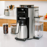A sleek, modern DeLonghi TrueBrew Fully Automatic Drip Coffee Machine CAM51035M in stainless steel with a thermal carafe sits on a kitchen counter. It features a digital display showing 7:30, multiple control buttons, a silver thermal carafe, and a transparent water reservoir. Nearby are jars filled with coffee beans and some kitchen utensils.