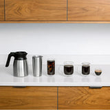 A lineup of coffee vessels on a white countertop in front of wooden cabinets. From left to right: a stainless steel thermal carafe, a stainless steel travel mug, a glass of iced coffee, a glass mug of black coffee courtesy of the DeLonghi TrueBrew Fully Automatic Drip Coffee Machine CAM51035M (Stainless Steel with Thermal Carafe), a smaller glass mug of black coffee, and an espresso cup.