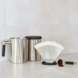 A sleek kitchen counter setup showcases a stainless steel carafe on the left, a cylindrical canister in the center, and the Moccamaster Technivorm Moccamaster Stainless Steel Coffee Filter Holder MA003 for cone-shaped filters on the right. In front, a measuring spoon with coffee grounds sits on the counter, keeping coffee filters easily accessible.