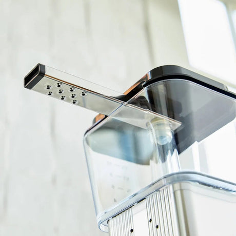 Close-up of a sleek, modern Moccamaster Technivorm Moccamaster 9-Hole Outlet Arm 53018 focusing on its transparent water reservoir and stainless steel filter holder. The machine has a minimalist design with clean lines and a glossy finish, positioned in a bright, well-lit environment.