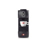 A black Illy Coffee Machine with E.S.E. pods (Illy ESE) has a small white ceramic cup positioned under the coffee spout. Both the cup and machine feature the Illy logo in red. The machine is compatible with illy E.S.E. pods and has two round buttons on the top front panel.