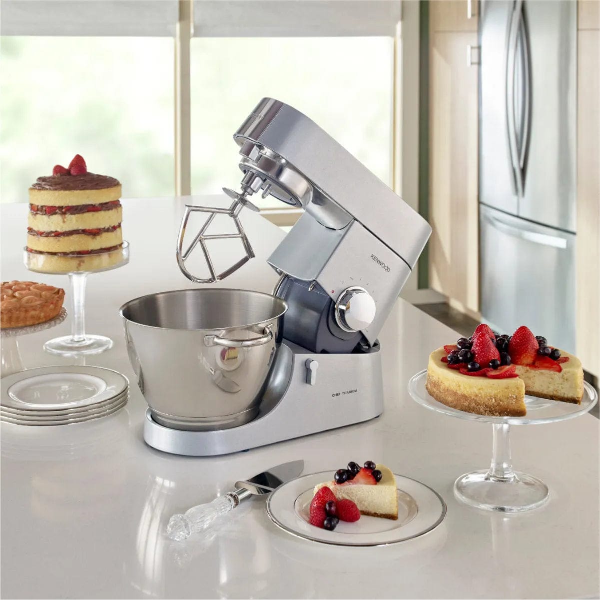 A Kenwood Chef Titanium KMC011 Stand Mixer & Kitchen Machine (4.7L / 5.0QT) sits on a kitchen counter, its 720 Watt motor ready for action. Nearby, two cakes on glass cake stands await serving; one topped with mixed berries and the other layered with chocolate icing. Plates and a cake server complete the scene.