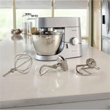 A Kenwood Chef Titanium KMC011 Stand Mixer & Kitchen Machine with a 720 Watt motor sits on a modern kitchen counter. The 4.7L / 5.0QT mixer bowl is attached, and three expandable attachments (a whisk, dough hook, and beater) are displayed in front. In the background, eggs in a bowl and jars decorate the vibrant countertop near a window.