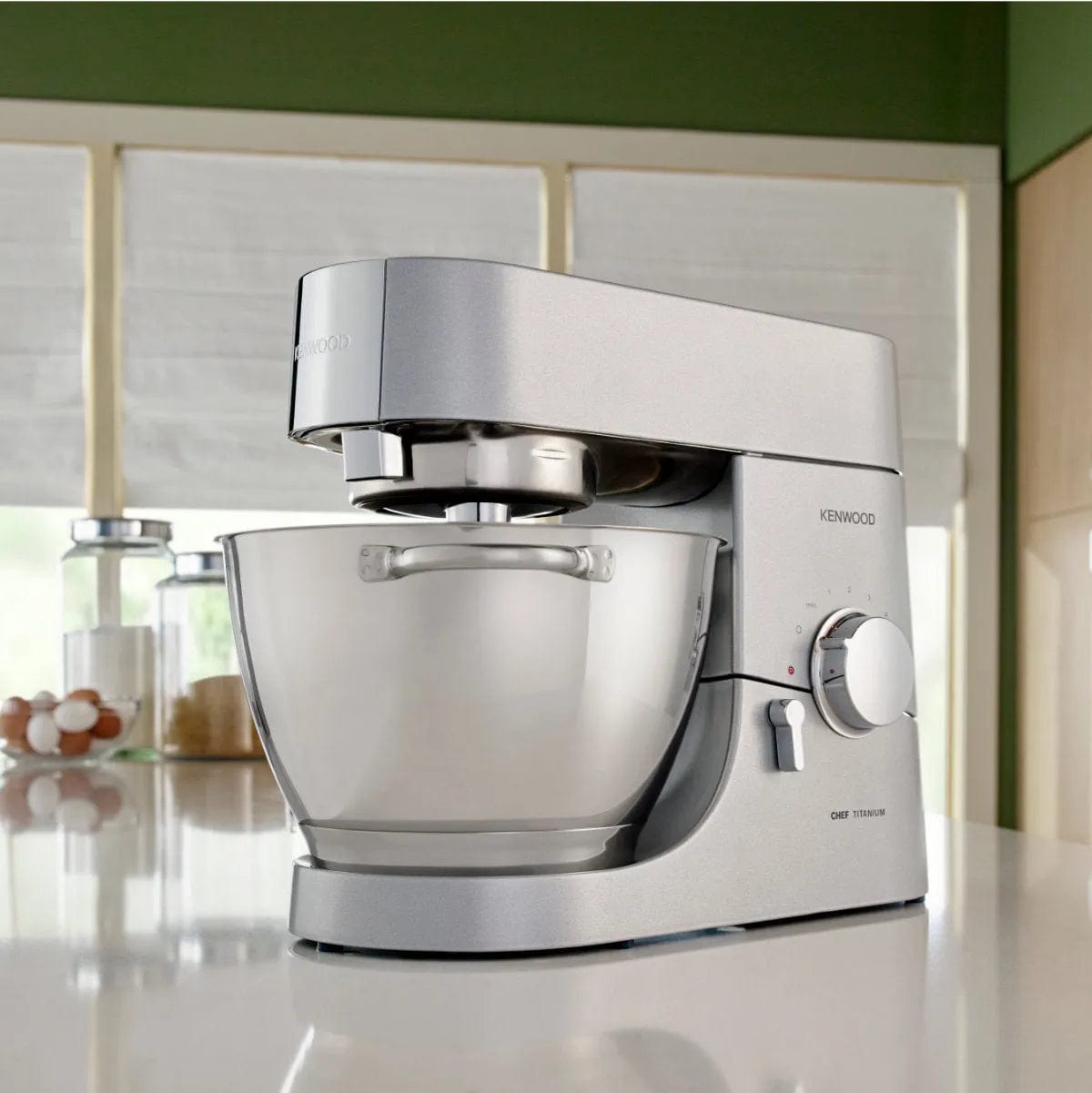 A Kenwood Chef Titanium KMC011 Stand Mixer & Kitchen Machine, boasting a powerful 720 Watt motor and featuring a large 4.7L / 5.0QT clear mixing bowl, is placed on the kitchen counter. The mixer is positioned in front of a window with white blinds, with various kitchen items including jars and eggs visible in the background.
