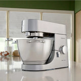 A Kenwood Chef Titanium KMC011 Stand Mixer & Kitchen Machine, boasting a powerful 720 Watt motor and featuring a large 4.7L / 5.0QT clear mixing bowl, is placed on the kitchen counter. The mixer is positioned in front of a window with white blinds, with various kitchen items including jars and eggs visible in the background.