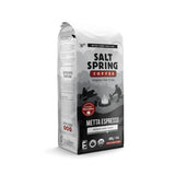 A 14-ounce bag of Salt Spring Metta Espresso Whole Bean Coffee sits against a white background. The packaging features a mountain landscape graphic and notes that the coffee is Organic Certified, Fair Trade Certified, and medium dark roast.