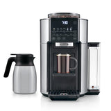 The DeLonghi TrueBrew Fully Automatic Drip Coffee Machine CAM51035M in stainless steel with a thermal carafe showcases a modern, sleek design. Its digital display reads "7:30" while brewing a mug of single-serve drip coffee. To the left of the machine sits the stainless steel carafe. The device features various buttons and a transparent water reservoir on its side.
