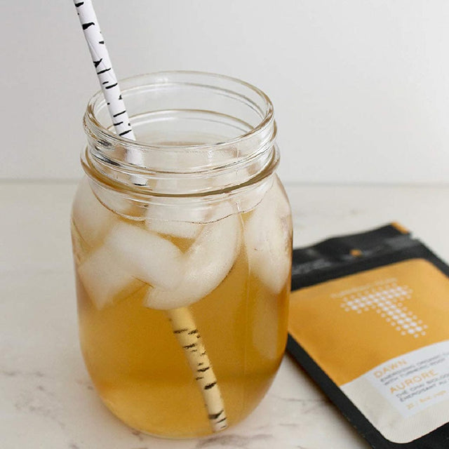 A mason jar filled with iced tea and ice cubes sits on a white surface. The drink, made from Turmeric Teas Dawn Black Chai Loose Leaf Tea, is stirred by a black and white striped paper straw. In the background, there is a yellow and black packet labeled with a large "T" from Turmeric Teas.