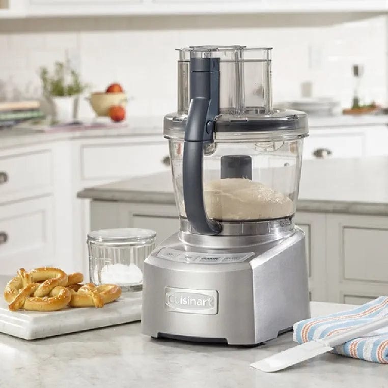 A Cuisinart® Elite Collection® 14-Cup (3.5 L) Food Processor FP-14DCNC rests on the kitchen countertop, dough inside its bowl. Accompanying it are a small cutting board with pretzels, a jar of flour, and a striped kitchen towel. The adjustable slicing disc enhances its high performance in this stylish, modern kitchen setting.